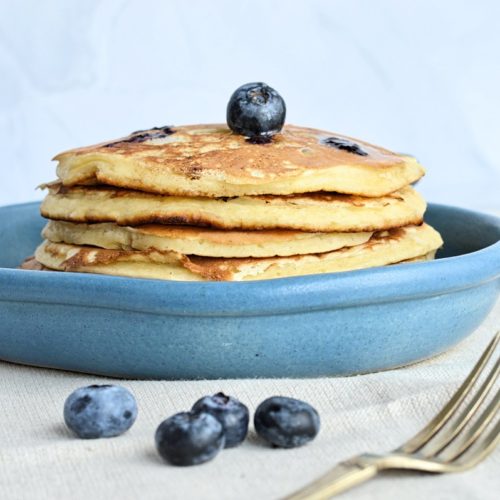 Pancakes aux bleuets dans assiette bleue avec fourchette et bleuets décoratifs.