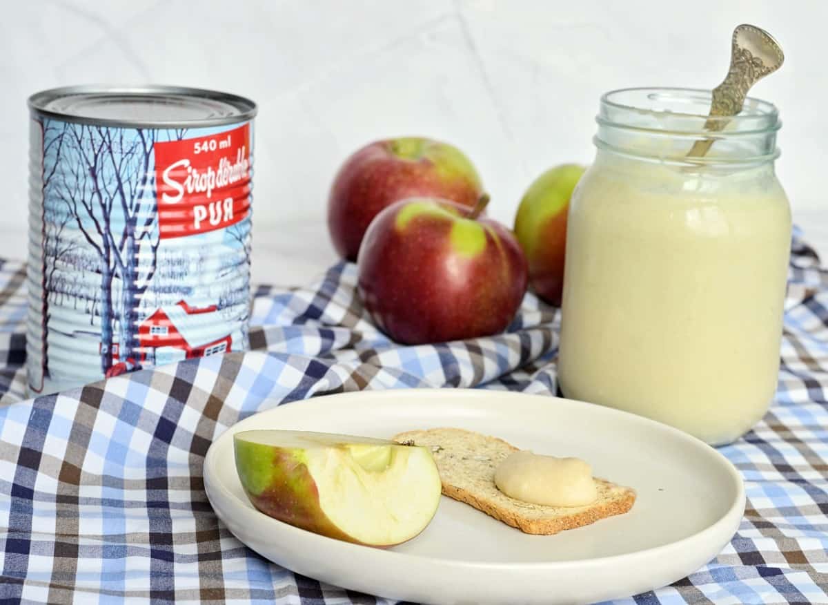 Un pot de beurre de pomme, 3 pommes derrière, une canne de sirop d'érable, une toast melba sur un assiette avec un peu de beurre de pomme.