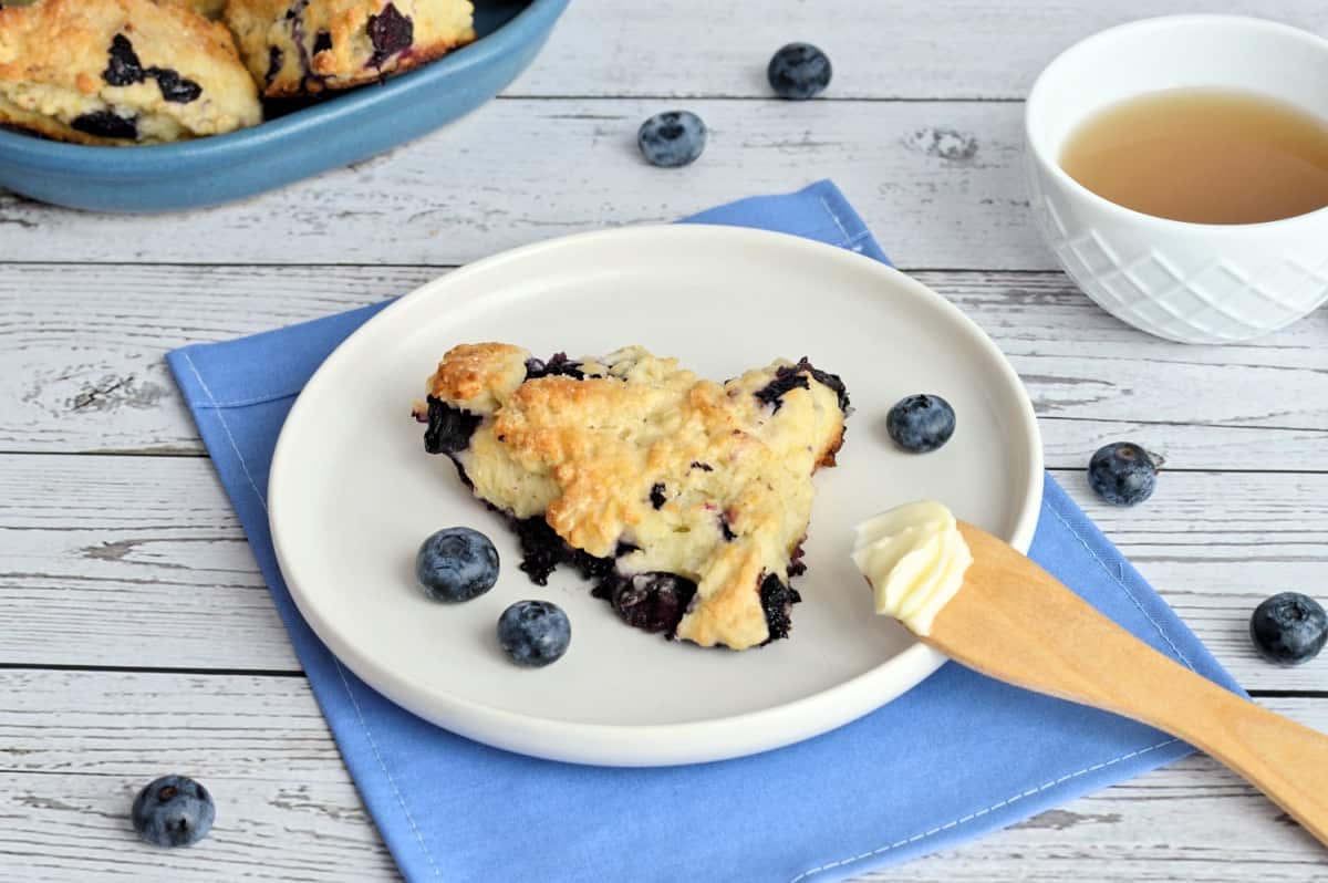 Un scone dans une assiette, un couteau avec du beurre, un bol de thé, une assiette remplie de scones aux bleuets derrière.