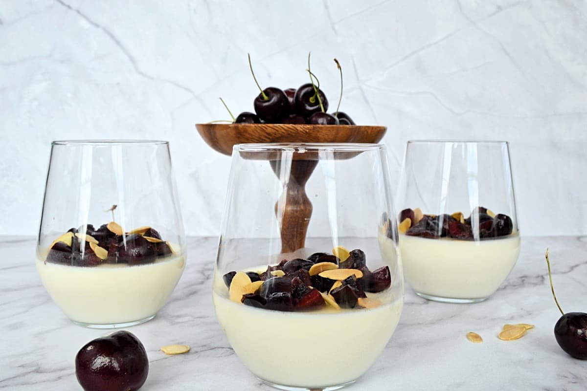 3 bols de panna cotta garnis de cerises et amandes, et des cerises fraîches autour.